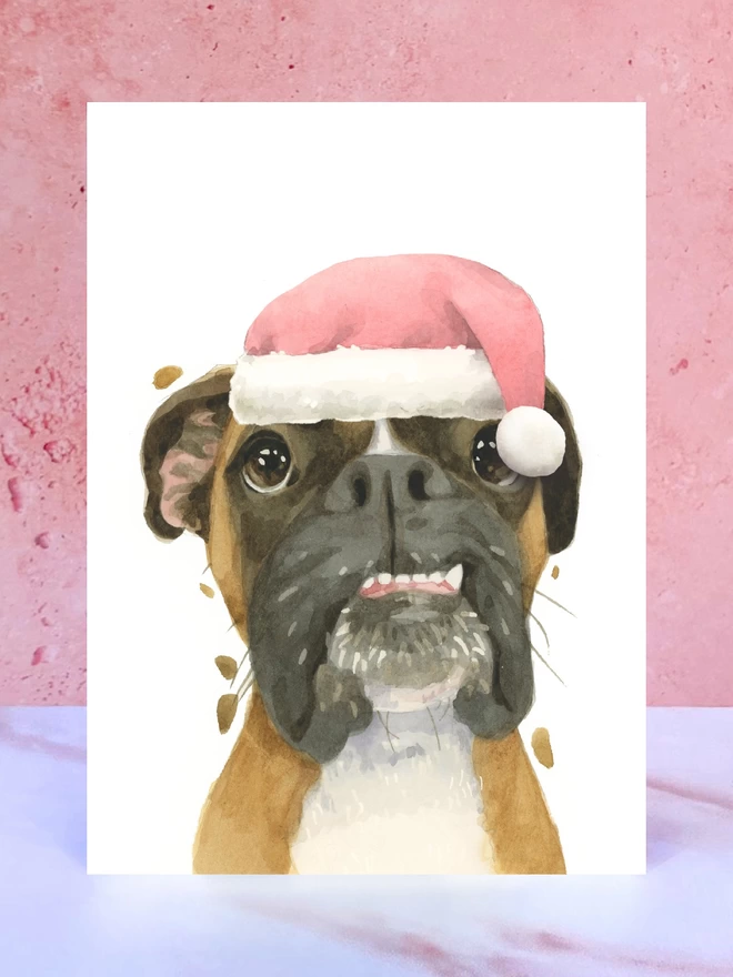 A Christmas card featuring a hand painted design of a boxer dog, stood upright on a marble surface surrounded by pompoms. 