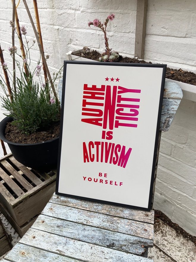 the print sits on an ageinFramed multicoloured typographic print of the quote “Authenticity Is Activism”  The print sits on an ageing garden chair, potted plants against a white wall.