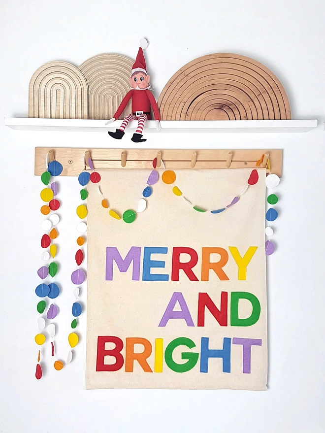 A cheerful image of the naughty elf sat on a shelf, below a banner hangs with Merry and Bright stitched in bright rainbow colours