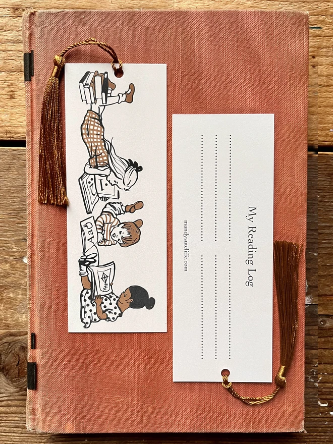 a photograph of a traditional card bookmark illustrated with a vintage children's book style drawing of kids reading books. The bookmark is cream, ochre and sepia in colour, the tassel is a golden brown. Laid on a kraft envelope and there is also a gift giving label, or bookplate sticker making it a lovely alternative to a greeting card