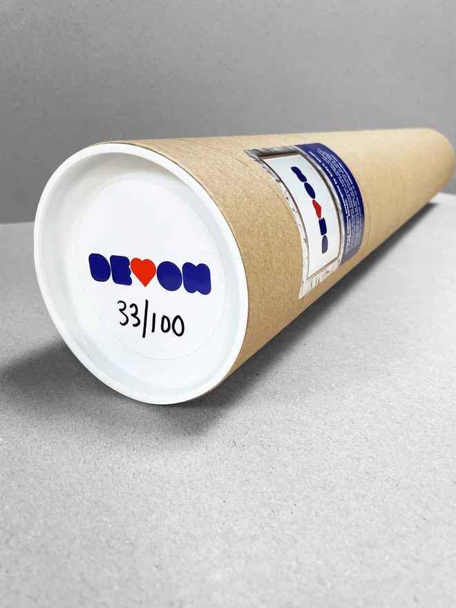 A cardboard tube laid on a plain grey background, containing the blue and red Devon print with a sticker on the end depicting that.