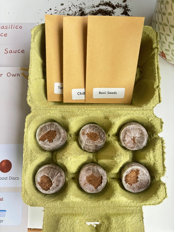 open green egg box with six compost discs inside. Inside the lid are three seed packets and instructions next to them.