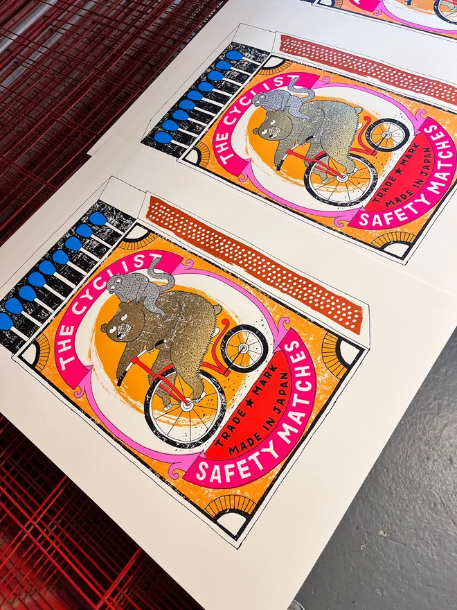Bear On A Bike Matchbox Screenprint Big Cyclist's Burden, a colourful matchbox design in the centre of a white screenprint. It sits next to another matchbox design on a studio rack. 