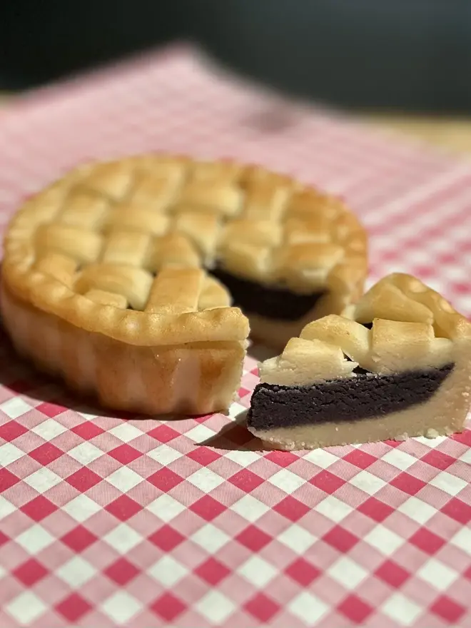Marzipan Cherry Pie, marzipan cherry pie on a traditional picnic cloth. It has been cut and a slice rests next to the whole pie. 