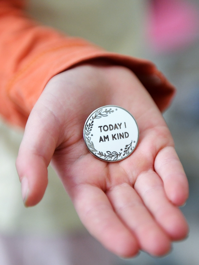 A round white enamel pin with a floral design and the words "Today I Am Kind" rests on the palm of a hand.