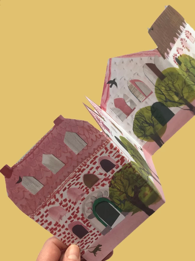 Hand opens up folded concertina card showing illustrated pink houses and green trees