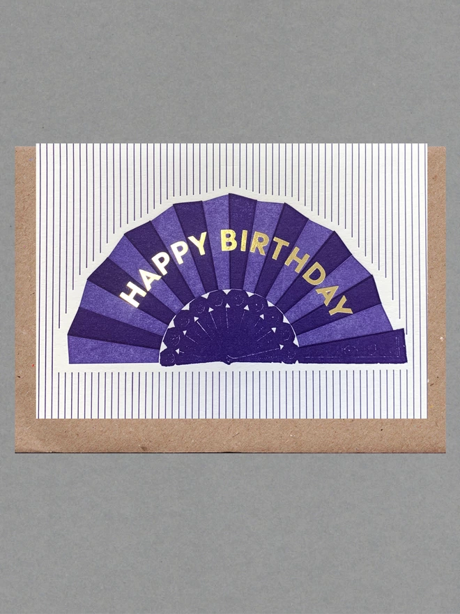 White card with purple fan with gold text reading 'Happy Birthday' on purple striped background with brown envelope behind