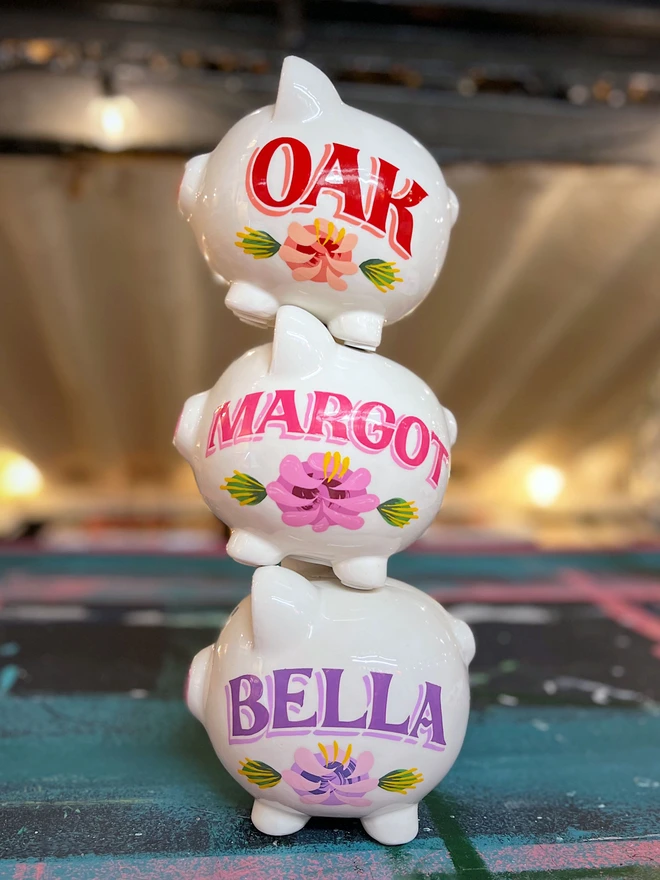 Three hand-painted white piggy banks stacked together. They are lettered with 'Oak', 'Margot' and 'Bella'. Oak is painted in red with a coral flower below it. Margot is painted in bright pink with a pink-purple flower below it. Bella is painted in bright purple with a purple flower below it.