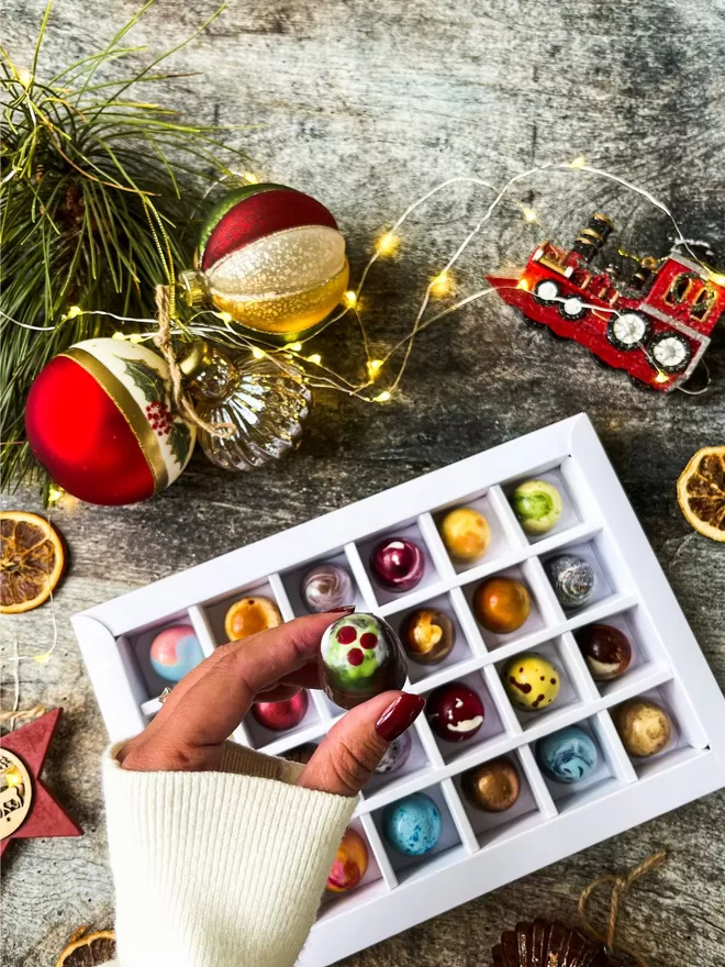 a hand holding a chocolate with a christmas pudding design in front of a box of colourful chocolates on a wood background surrounded by christmas decorations and lights