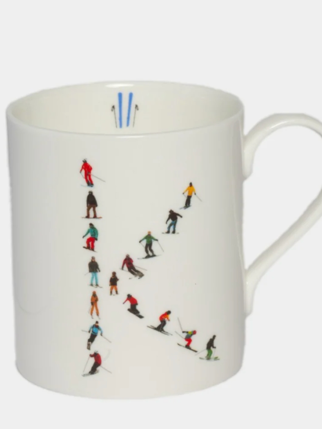 A simple white mug illustrated with a ‘K’ made of small, multicoloured skiers. The mug is set against a plain, white background.