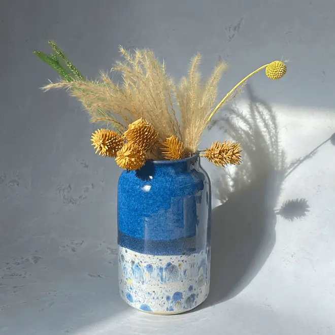 Shoreline Large Vase, a colourful vase placed on a stone surface in front of a plain backdrop. It is accompanied by flowers. 