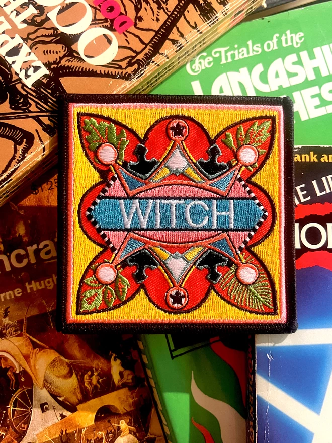 The patch sits on old fashioned books, at the centre of the patch is the word “Witch” is embroidered in white on a light blue strip. Surrounding this is a colourful, bold design which sits on a yellow background, with a thin pink inner border and a thick black outer border.