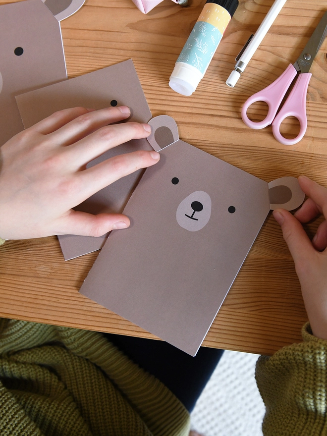 Several brown bear greetings cards lay on a wooden table. A child is gluing the ears to the corners of one card.