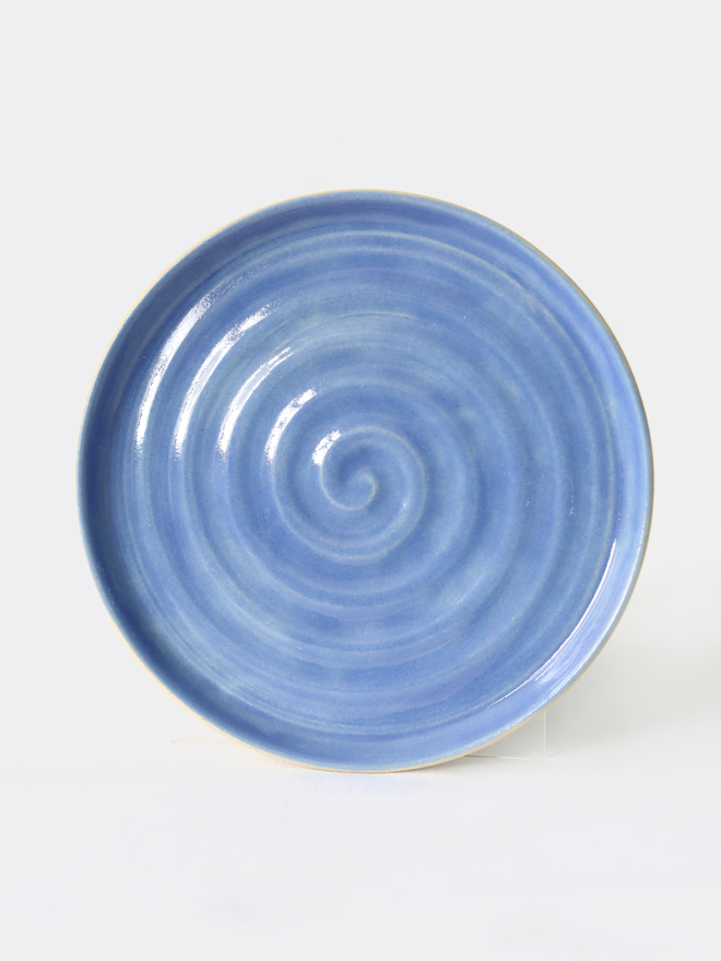 A handmade ceramic cake plate in pacific blue glaze, photographed from the front against a plain light grey background, showing a natural concentric swirl texture and a shallow raised rim