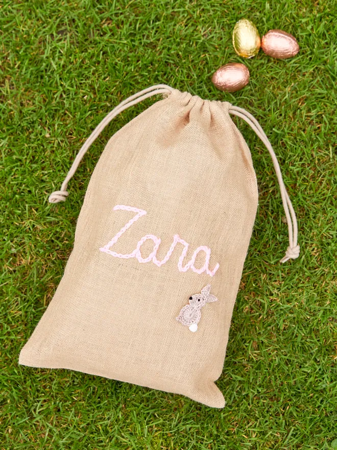 jute drawstring sack with brown knitted bunny decoration and pink 'zara' name next to golden chocolate eggs on green grass
