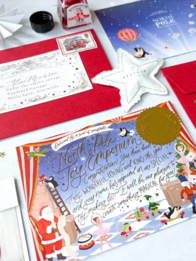 An angled view of the illustrated "North Pole Toy Emporium" notecard with a gold foil seal and the message "Congratulations! You have been truly wonderful, loving and kind this year". Behind it are a red envelope with a personalised label and vintage stamp, the starry blue card back and a felt "Believe" star.