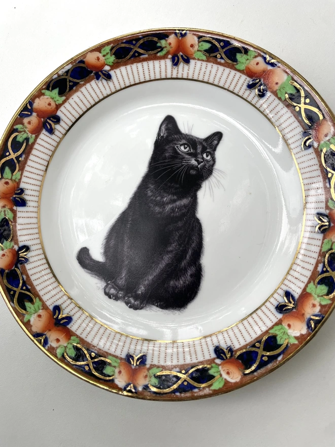 a vintage plate with a dark red border with intricate gold patterning, in the centre of the plate is a vintage style black and white illustration of a black witch's cat