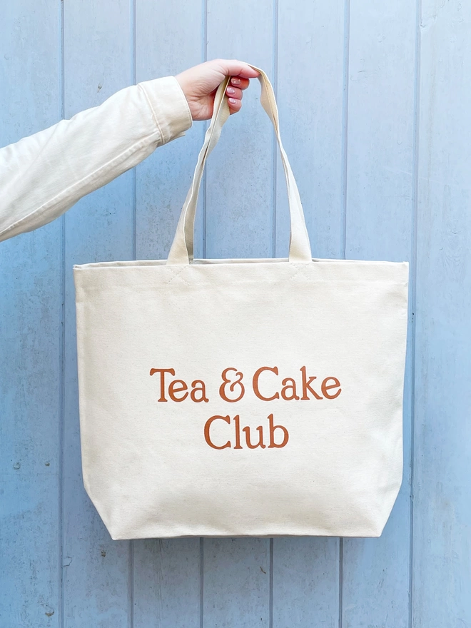 model holding a large natural canvas tote bag bearing the words tea and cake club