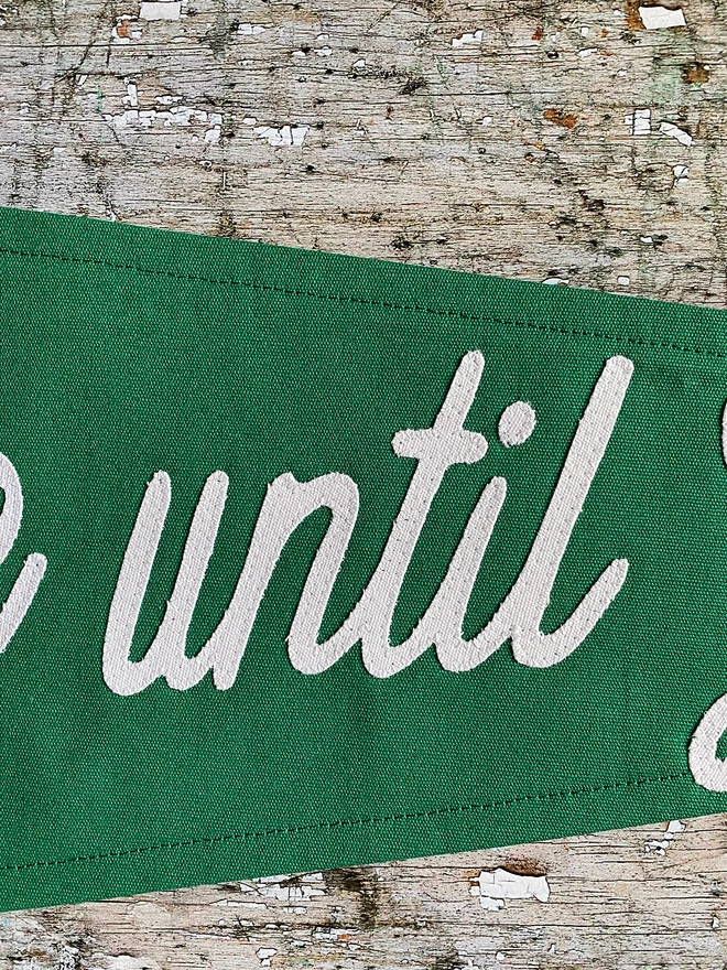 A close up detail of the word until written in ivory canvas. Detail of a green Coffee before Gin pennant flag.