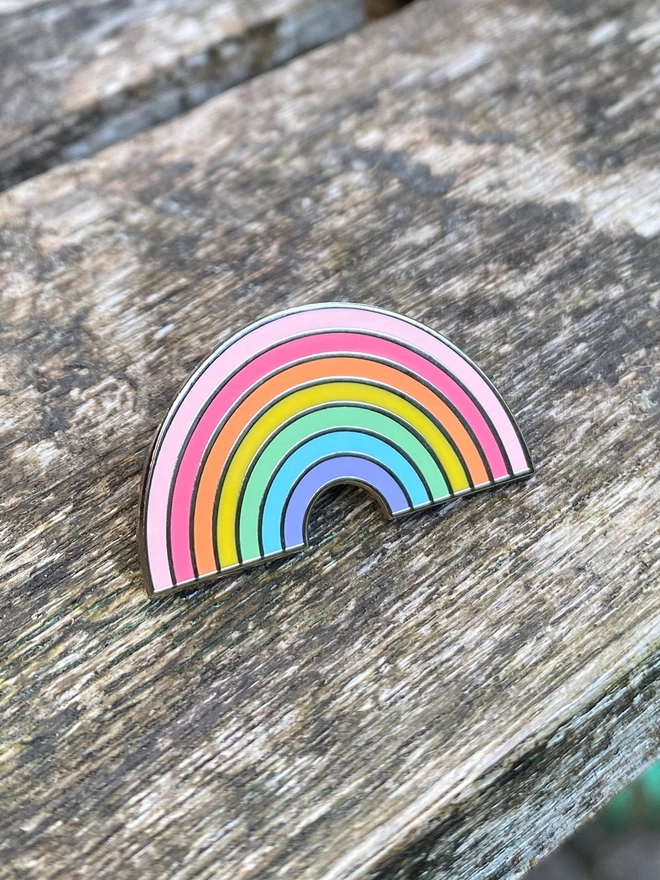 A small pastel rainbow enamel pin badge is on a wooden table.