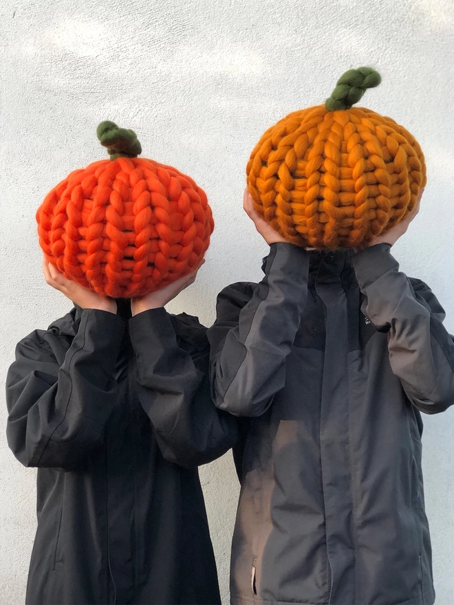 Two people holding up hand knitted pumpkins seen in front of their head.