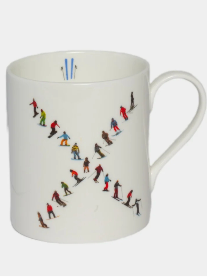 A simple white mug illustrated with an ‘X’ made of small, multicoloured skiers. The mug is set against a plain, white background.
