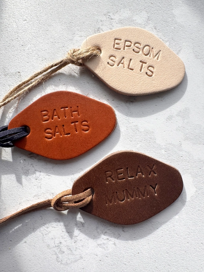 "Three hand-stamped leather tags in brown, cream, and tan, with the words 'RELAX MUMMY,' 'EPSOM SALTS,' and 'BATH SALTS' embossed. Each tag has a cord for tying around jars, displayed on a light, textured surface.