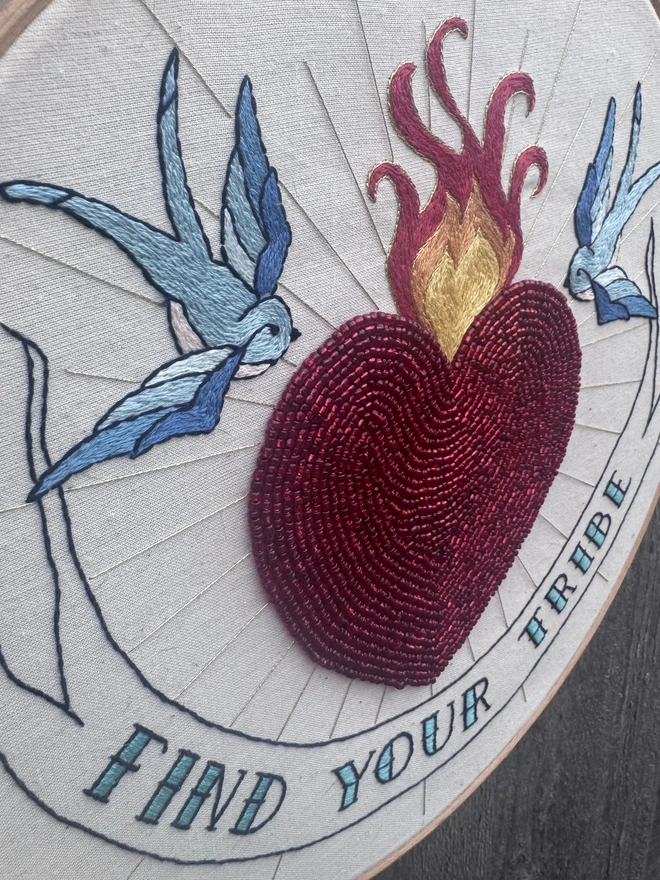 Embroidery hoop containing a red beaded heart with fire gold rays two bluebirds and a banner containing the words find your tribe 