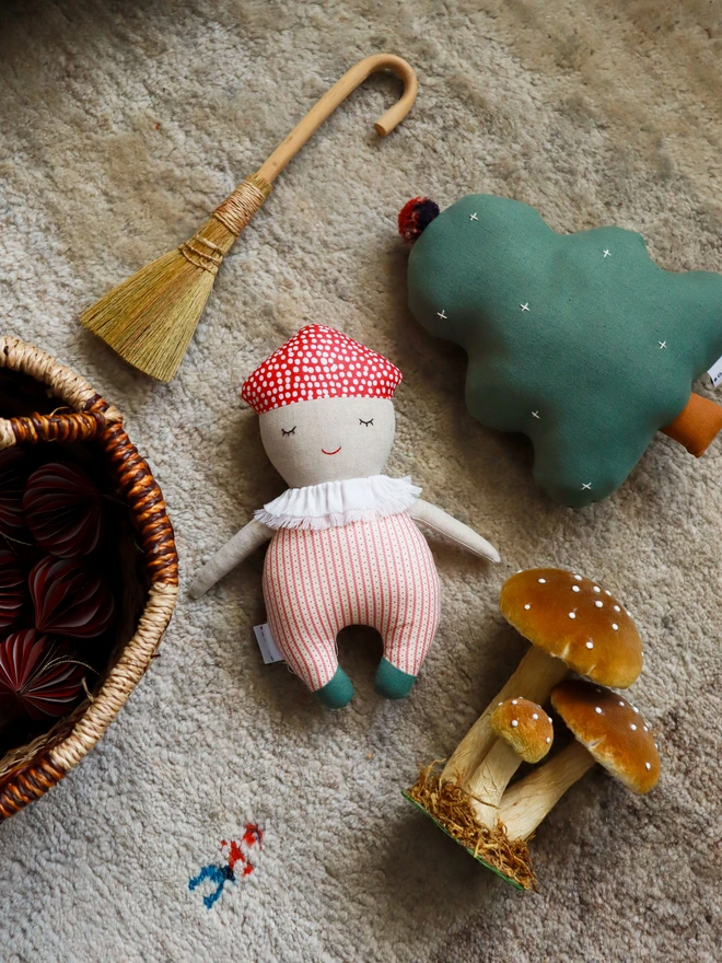 Soft handmade mushroom-themed doll with a red spotted cap, embroidered smiling face, ruffled white collar, red-striped body, and small green feet.