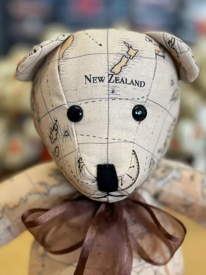Teddy bear made from map-print fabric sitting upright on a wooden table, wearing a brown bow.