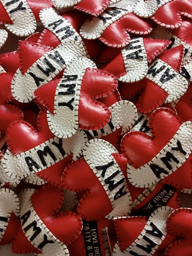 A pile of red faux leather padded heart brooches, with AMY in black lettering on white scrolls