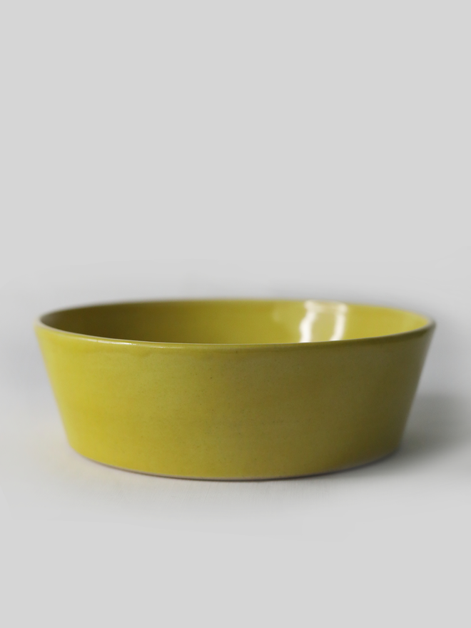A single wide, shallow glazed ceramic pasta bowl in sun yellow, photographed from a slight angle against a plain light grey background, showing the unglazed natural clay base.