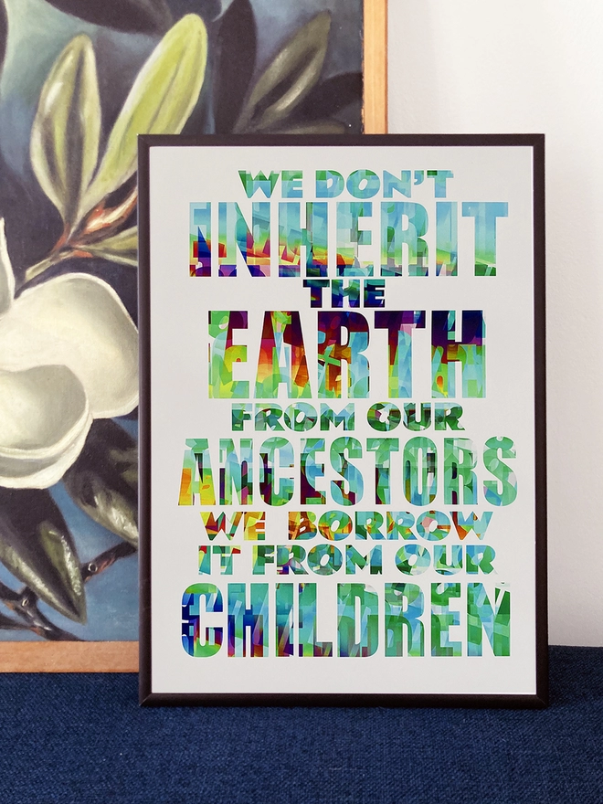 Framed multicoloured typographic print of a Pulp song lyric from Common People - “We don't inherit the earth from our ancestors, we borrow it from our children”. The prints rests against a painting of white flowers with a blue background.