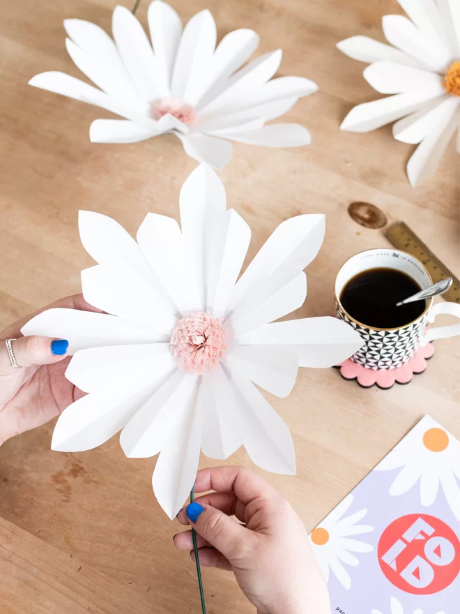 Woman's hands holding a giant paper daisy, with more finished paper daisies, cup of coffee and varies tools on a table.