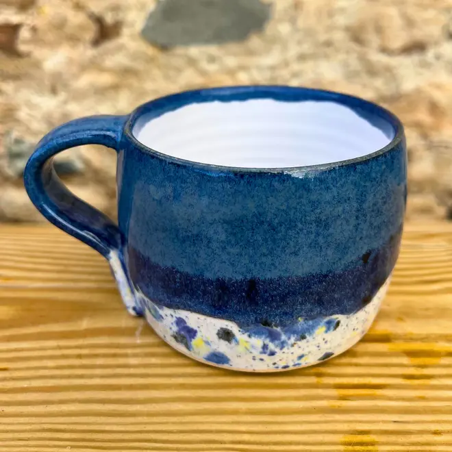Shoreline Large Round Mug, a colourful mug on a wooden surface against a brick backdrop. 