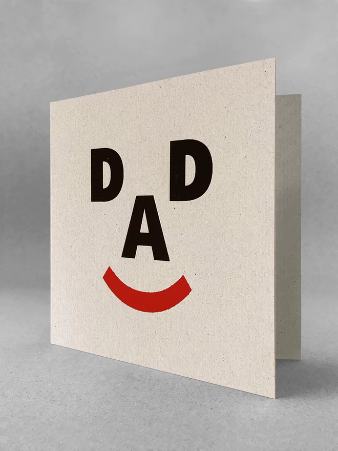 The letters D, A, D and a closed bracket symbol make up a happy face, screenprinted on recycled grey card. Stood slightly open, in a light grey studio set