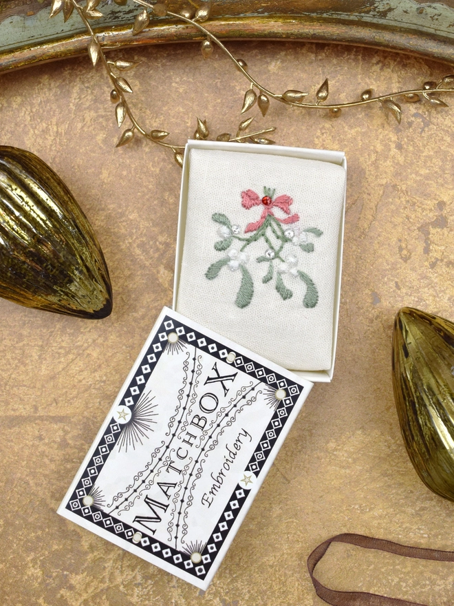A sprig of mistletoe, inside a Matchbox displayed on a gold background with glass baubles.