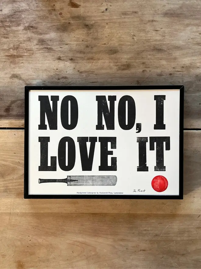 Letterpress of 'No No, I Love It' with a cricket bat and cricket ball, framed with a black frame on a wooden table