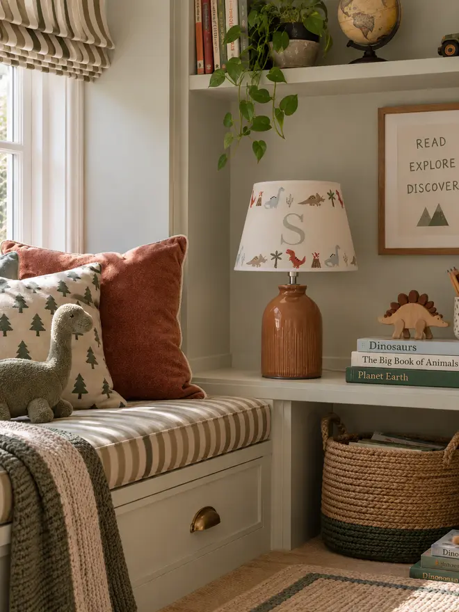 Bright children’s reading nook with personalised dinosaur lampshade, dinosaur toys, books and a striped window seat in a boy’s bedroom.