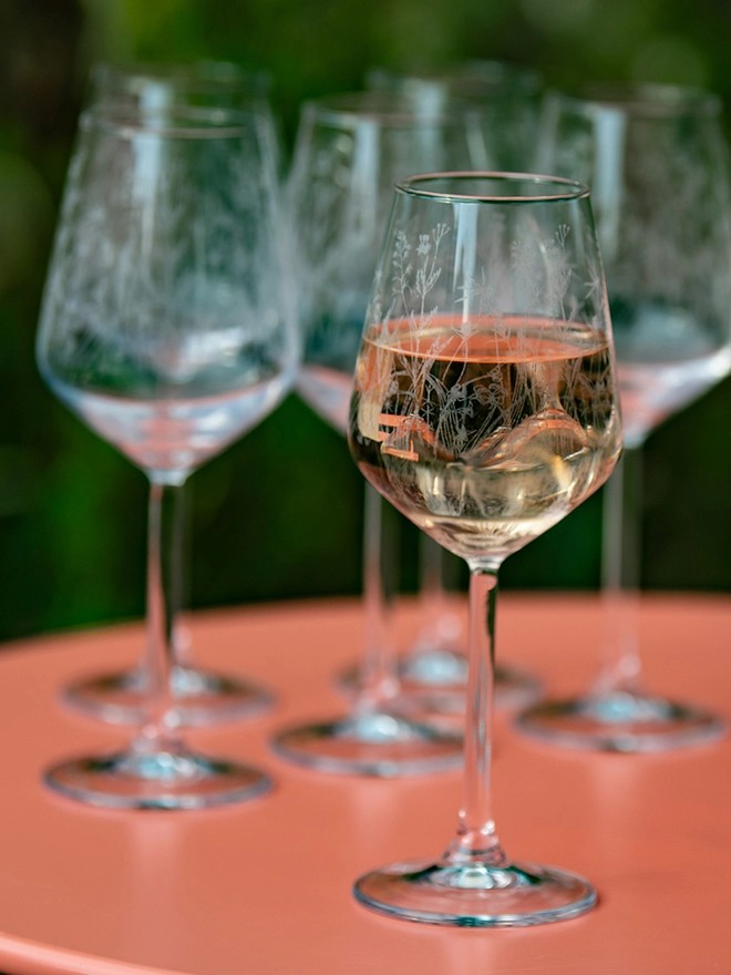 Meadow Wine Glass, a group of six etched wine glasses with wildflower patterns sit on a bright pink patio table. 