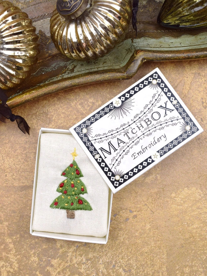 A bright green Christmas tree, inside a Matchbox displayed on a gold background with glass baubles.