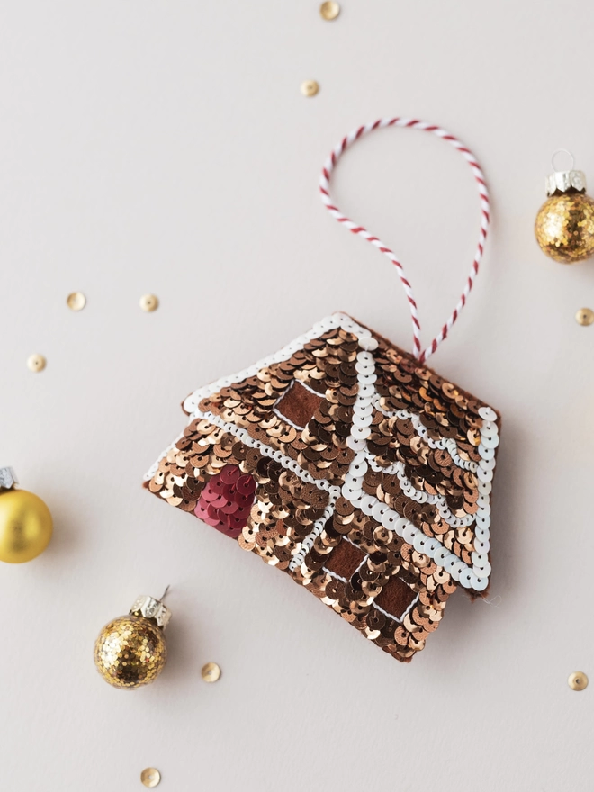 A sequinned gingerbread house ornament on a cream table surrounded by mini baubles