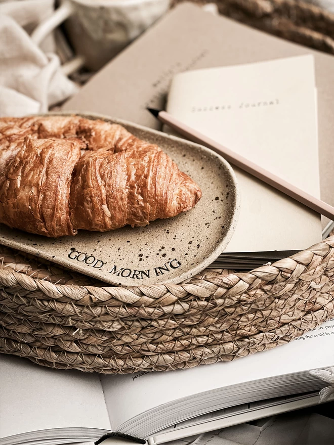 croissant on an oval, beige speckled plate with the words 'good morning' stamped in black around the edge