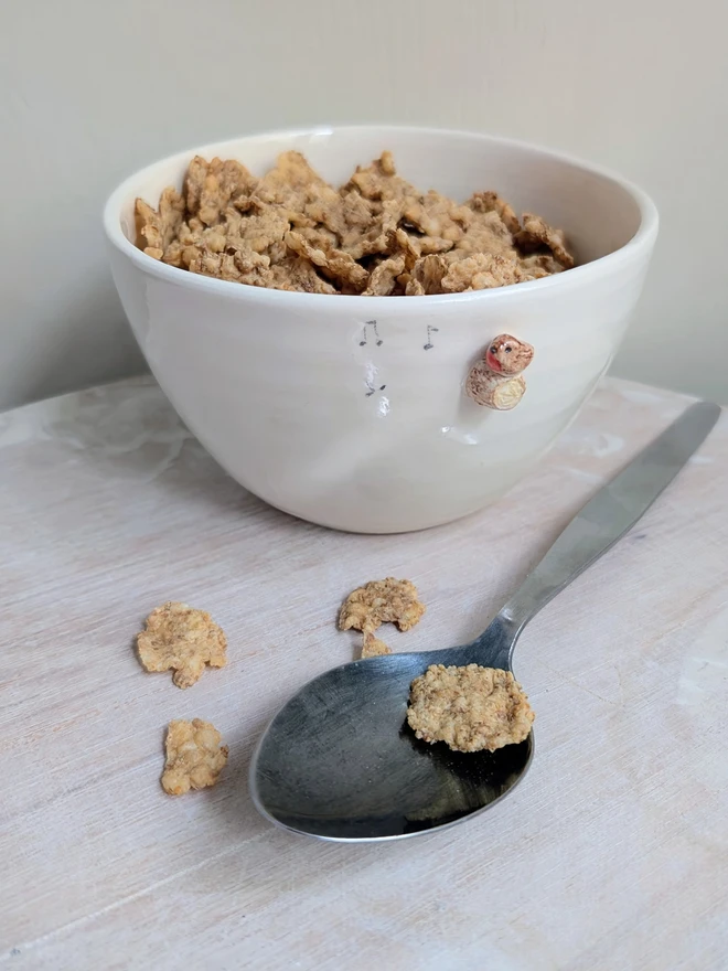 white ceramic bowl with a tiny modelled robin perching on a modelled branch with musical notes, the bowl is full of breakfast cereal and there is a spoon resting beside it