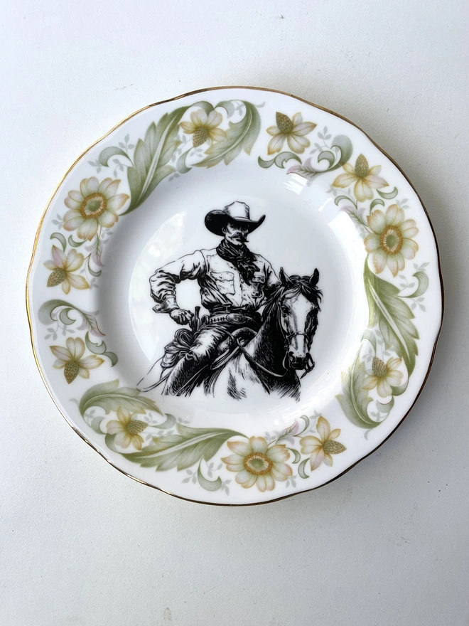 vintage plate hanging on a white wall, it has a decorative border, with a printed vintage illustration of a cowboy in the middle 