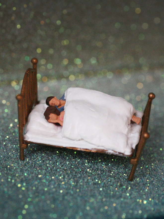 Miniature scene in an artbox showing a tiny man and woman lying in bed against a sparkling backdrop evoking the night sky. 