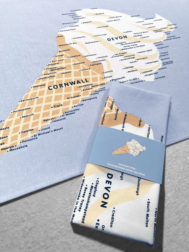 A folded teatowel in a wrap lies on top of an open tea towel, both with the Devon and Cornwall Ice Cream Map design, both sat on a grey background.