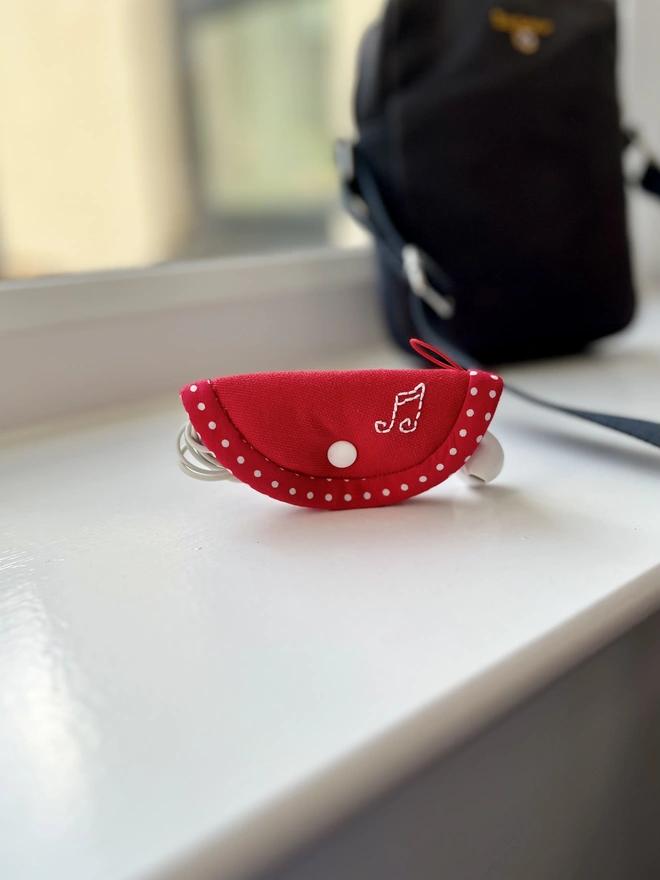 Small soft cotton red earphone case embroidered with musical notes on windowsill, crossbody bag in background.