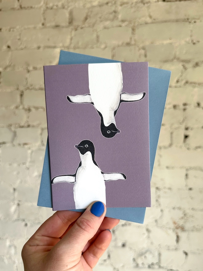 a purple greetings card featuring two illustrations of a penguin with a blue envelope