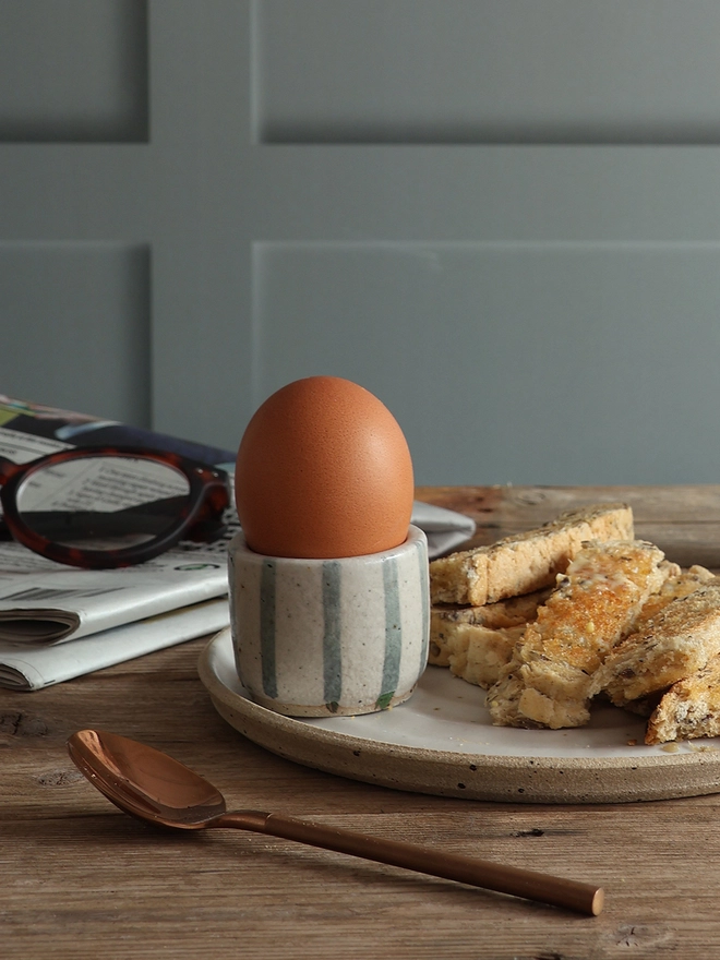 A breakfast scene featuring a handmade ceramic egg cups with a blue and white stripe design holding a boiled egg. The egg cup sits on a white handmade ceramic plate filled with buttered toast. A brass spoon and a newspaper with a pair of glasses on top also sit on the table.  